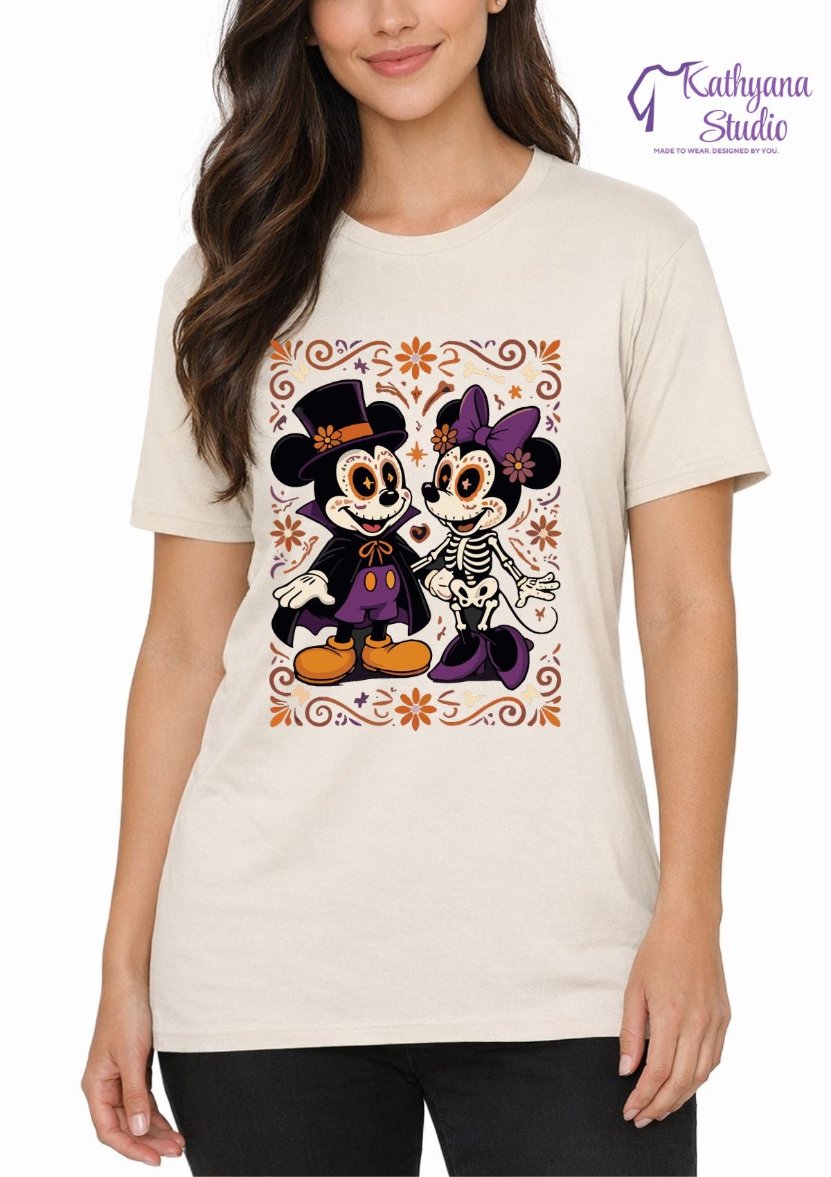 A woman wearing a cream-colored t-shirt with a colorful graphic of Mickey and Minnie Mouse styled as Day of the Dead skeletons, on a white background with a 'Kathyana Studio' watermark.